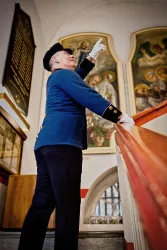 Ein Mann in einer uniformierten blau-grauen Jacke steht an einer Treppe und weist auf etwas hin. Im Hintergrund sind religiöse Gemälde an der Wand sichtbar.