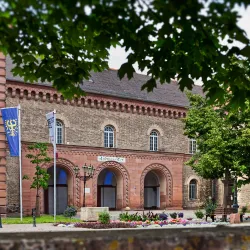 Ein historisches Gebäude mit roten Backsteinmauern und schönen Fenstern. Darum herum sind grüne Bäume und bunte Blumen.