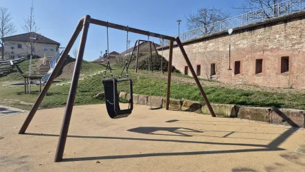 Ein Spielplatz mit einer großen Schaukel auf einem sandigen Boden. Im Hintergrund sind grüne Rasenflächen und eine historische Mauer zu sehen.