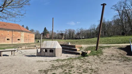 Ein Spielplatz mit einer Holzstruktur und einer Sandfläche. Im Hintergrund sind Bäume und eine Wiese sichtbar.