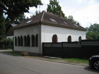 Ein zweigeschossiges Haus mit einem roten Ziegeldach und weißen Wänden. Es hat mehrere Fenster mit Fensterläden und ist von einem schwarzen Zaun umgeben.