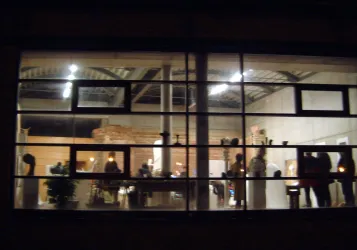 Eine moderne Ausstellung in einem gut beleuchteten Raum. Menschen stehen und betrachten die Kunstwerke durch große Fenster.