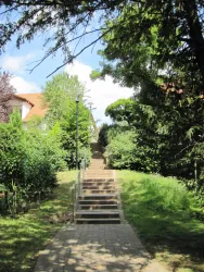 Ein Weg mit Treppen führt durch eine grüne Umgebung. Im Hintergrund sind Bäume und Gebäude sichtbar.