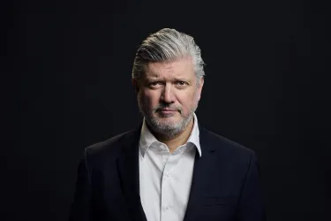 Ein reifer Mann mit grauen Haaren und einem Bart steht vor einem dunklen Hintergrund. Er trägt ein schwarzes Jackett und ein weißes Hemd.