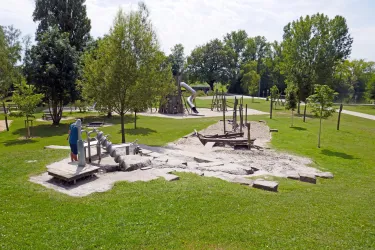 Ein Spielplatz mit verschiedenen Spielgeräten und einer Rutsche ist in einem grünen Park zu sehen. Bäume und Wiesen umgeben den Bereich, der für Kinder zum Spielen einlädt.