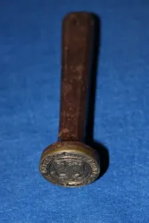 Ein leichter Holzstempel mit einem runden, metallischen Kopf. Die Oberseite zeigt ein geprägtes Symbol oder eine Figur.