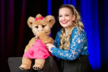 Eine lächelnde Frau präsentiert eine Stofftierpuppe in einem pinken Kleid. Im Hintergrund sind bunte Lichter zu sehen.
