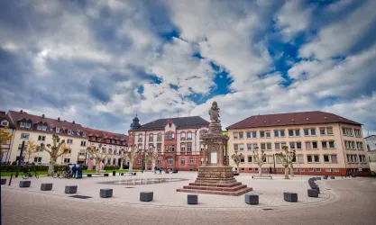 Ein schöner Platz mit einem Denkmal in der Mitte und mehreren Gebäuden im Hintergrund. Der Himmel ist bewölkt mit einigen blauen Stellen.