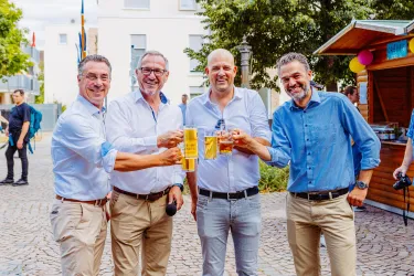 Vier Männer stoßen mit Bier in der Hand an, sie lächeln und scheinen eine gute Zeit zu haben. Im Hintergrund ist eine Stadtansicht mit Bäumen und festlicher Atmosphäre zu sehen.