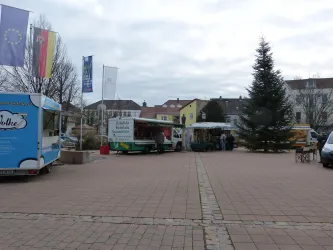 Ein Platz mit mehreren Food Trucks und einem Weihnachtsbaum. Im Hintergrund sind bunte Gebäude und Fahnen zu sehen.
