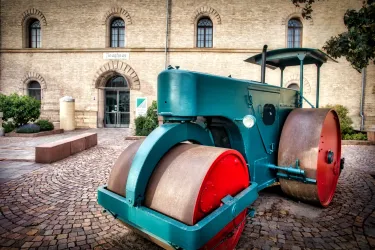 Ein alter grüner Straßenwalze steht auf einem gepflasterten Platz vor einem historischen Gebäude. Die Walze hat rot lackierte Rollen und zieht die Aufmerksamkeit auf sich.