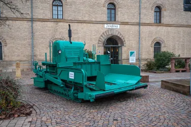 Ein grüner Schnee-räumtraktor steht vor einem historischen Gebäude. Die Umgebung ist gepflastert und es sind einige Pflanzen zu sehen.