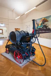 Eine historische Dampflokomotive steht in einem Museum. Sie ist schwarz mit roten Rädern und befindet sich auf einem Holzplattform.