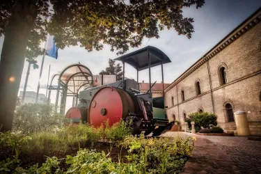 Ein historischer Straßenwalzen mit roten Walzen steht vor einem alten Gebäude. Im Hintergrund sind einige Bäume und Flaggen zu sehen.