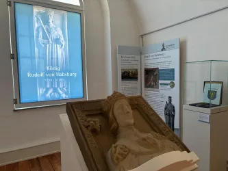 Eine Skulptur von König Rudolf von Habsburg steht in einem Museum. Im Hintergrund sind Informationsplakate und ein Fenster mit einer Darstellung des Königs zu sehen.