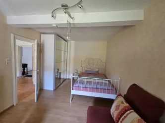 Ein helles Zimmer mit einem einzelnen Bett und einem roten Sofa. Der Raum hat einen freundlichen, modernen Stil und viel Platz.