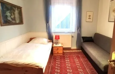 Ein einfaches Zimmer mit einem Einzelbett und einer Schlafcouch. Es gibt einen kleinen Tisch mit einer roten Lampe und ein Fenster mit lässigen Vorhängen.