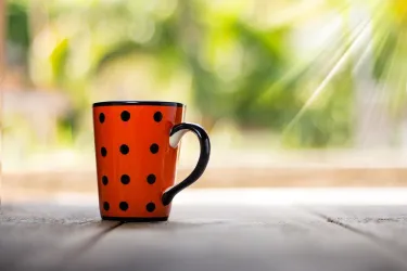 Eine orangefarbene Tasse mit schwarzen Punkten steht auf einem Holztisch. Im Hintergrund ist unscharf die grüne Natur zu sehen.