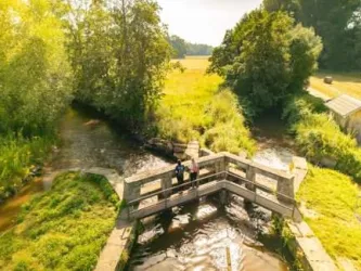 Eine Holzbrücke über einen kleinen Fluss, umgeben von grünen Bäumen und Wiesen. Im Hintergrund sind weitere Bäume und eine ländliche Landschaft zu sehen.