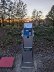 Eine Parkuhr steht auf einem gepflasterten Weg. Im Hintergrund sind Bäume und ein Sonnenuntergang sichtbar.