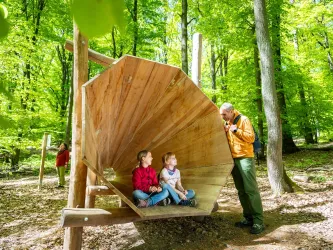 Zwei Kinder sitzen in einem großen Holzlautsprecher im Wald. Ein Erwachsener steht daneben und spricht mit ihnen.