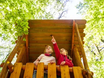 Zwei Kinder stehen auf einem Spielturm im Wald und schauen nach oben. Eines der Kinder zeigt mit dem Finger auf etwas im Baum.