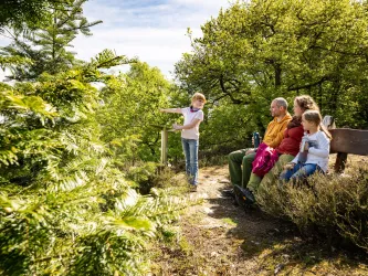 Eine Familie sitzt entspannt auf einer Bank in der Natur. Ein Kind zeigt auf etwas in der Ferne, umgeben von grünen Bäumen und Sträuchern.