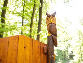 Eine geschnitzte Holz-Eule sitzt auf einem Pfosten im Wald. Der Hintergrund zeigt grüne Bäume und sanftes Licht.