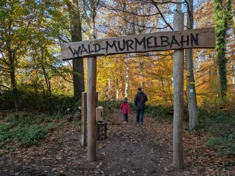 Ein Holzschild mit der Aufschrift "Wald-Murmelbahn" steht am Eingang eines Waldes. Im Hintergrund gehen ein Erwachsener und ein Kind auf einem Pfad durch die bunten Herbstbäume.