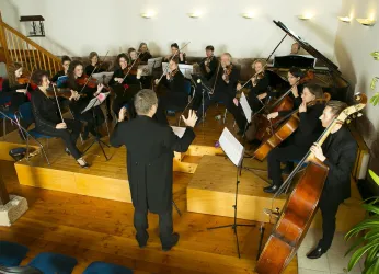 Ein Orchester spielt in einem gemütlichen Raum. Der Dirigent leitet die Musiker, die verschiedene Instrumente spielen.