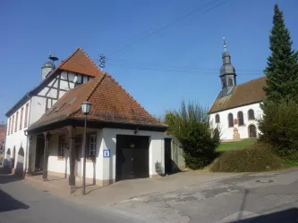 Ein malerisches Dorf mit einem traditionellen Fachwerkhaus und einer Kirche im Hintergrund. Der Himmel ist klar und blau.