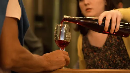 Eine Person schenkt Wein in ein Glas. Im Vordergrund ist ein Handgelenk zu sehen, während im Hintergrund eine andere Person schaut.