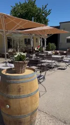 Eine gemütliche Terrasse mit Tischen und Stühlen unter Sonnenschirmen. Im Vordergrund steht ein Holzfass mit einer Blumenschale.