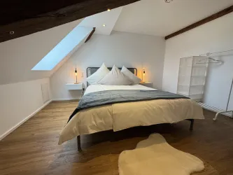 Ein helles, modernes Schlafzimmer mit einem großen Bett und weißer Bettwäsche. Auf dem Boden liegt ein weicher Teppich, und es gibt einen offenen Kleiderschrank.