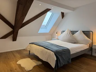 Ein modernes Schlafzimmer mit einem großen Fenster und einem bequemen Bett. Holzdetails und gemütliche Dekoration schaffen eine einladende Atmosphäre.