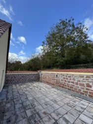 Eine leere Terrasse mit Steinfussboden und einer Mauer im Hintergrund. Der Himmel ist blau mit vereinzelten Wolken und es gibt Bäume in der Nähe.