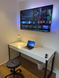 Ein moderner Arbeitsplatz mit einem Schreibtisch, einem Laptop und einem Monitor an der Wand. Im Hintergrund leuchtet eine kleine Lampe und es gibt einen Mülleimer.