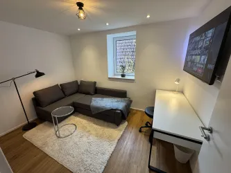 Ein modernes Wohnzimmer mit einem grauen Sofa, einem kleinen Tisch und einem Schreibtisch. Natürliches Licht fällt durch das Fenster und schafft eine einladende Atmosphäre. Schlafsofa, Arbeitszimmer, TV-Zimmer.