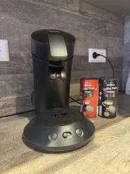 Eine schwarze Kaffeemaschine steht auf einem Tisch. Daneben befinden sich zwei Dosen mit Kaffeepads für klassischen Kaffee und Espresso.