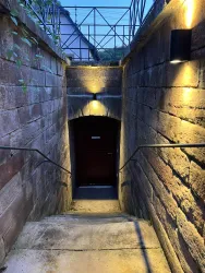 Ein dunkler Treppenabgang, der zu einer geschlossenen Tür führt. Der Bereich ist schwach beleuchtet und von einer steinernen Wand umgeben.