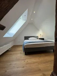 Ein helles Schlafzimmer mit schrägen Wänden und einem Dachfenster. Das Bett ist ordentlich gemacht und ein Holzfußboden verleiht dem Raum eine gemütliche Atmosphäre.
