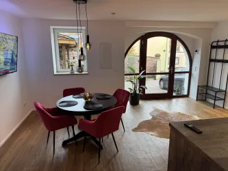 Ein modernes Wohnzimmer mit einem runden Tisch und pinken Stühlen. Durch das große Fenster und die Glastür fällt viel Licht hinein.