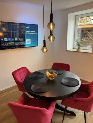 Ein modernes Esszimmer mit einem runden Tisch und pinken Stühlen. An der Wand hängt ein Fernseher, während eine Pflanze auf der Fensterbank steht.