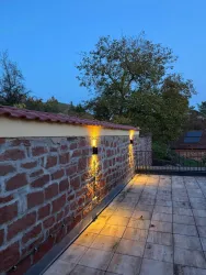 Eine Terrasse mit beleuchteter Steinmauer und Blick auf Bäume. Der Himmel ist in der Dämmerung.