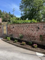 Eine gepflasterte Einfahrt mit einer roten Ziegelmauer. Im Vordergrund sind mehrere kleine Pflanzen und Blumen angeordnet.