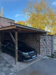 Ein schwarzes Auto steht unter einem überdachten Carport. Im Hintergrund sieht man herbstliche Bäume und einen blauen Himmel.