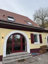 Ein charmantes Haus mit gelber Fassade und roten Fensterläden. Der Eingangsbereich ist einladend mit einem Sitzplatz neben der Tür.