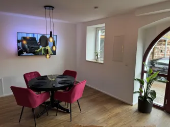 Ein modernes Esszimmer mit einem runden schwarzen Tisch und vier pinken Stühlen. An der Wand hängt ein Fernseher, und ein Fenster sowie eine Pflanze sorgen für ein helles Ambiente.