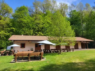 Ein gemütliches Gebäude in der Natur mit einer grünen Wiese und Bänken. Es sind Sonnenschirme aufgestellt, und Bäume umgeben den Bereich.