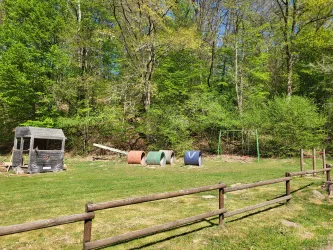 Ein Spielplatz in der Natur mit einer kleinen Holzhütte und bunten Tunnelröhren. Grüne Wiesen und Bäume umgeben den Bereich.